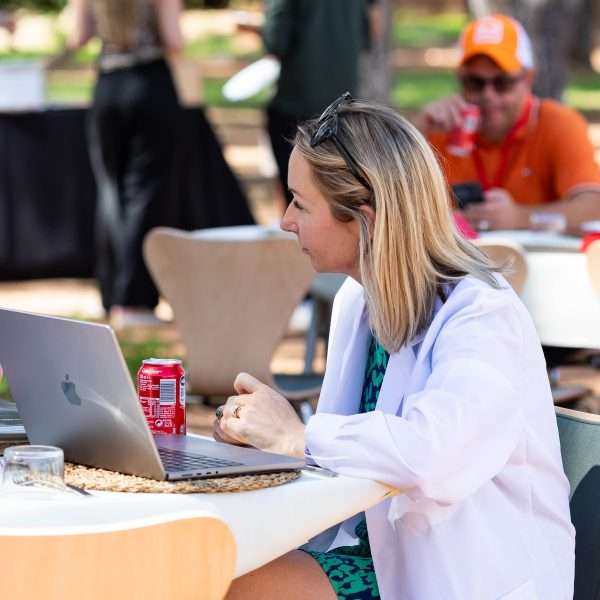 A woman in a white coat sits at an outdoor table, working on a laptop with a can of Coke nearby. Other people are seated and walking in the background in a sunny, casual setting.