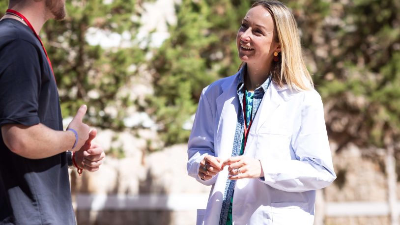 A man in sunglasses talks with Vicki Young in a white lab coat outdoors, green trees and sunlight behind them. Both appear to be smiling and engaged in conversation about a company’s recent rebrand done by Nalla