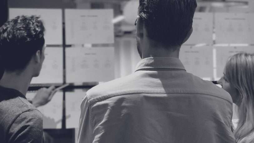 Three people viewed from behind look at papers or charts posted on a glass wall. One person gestures towards the papers as they discuss or review the content together in an office or workspace setting.