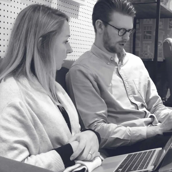 Two people sit side by side in an office, looking at a laptop together—a typical scene at a brand strategy agency. The woman faces the laptop, while the man, wearing glasses, looks at a tablet or document. The photo is in black and white.